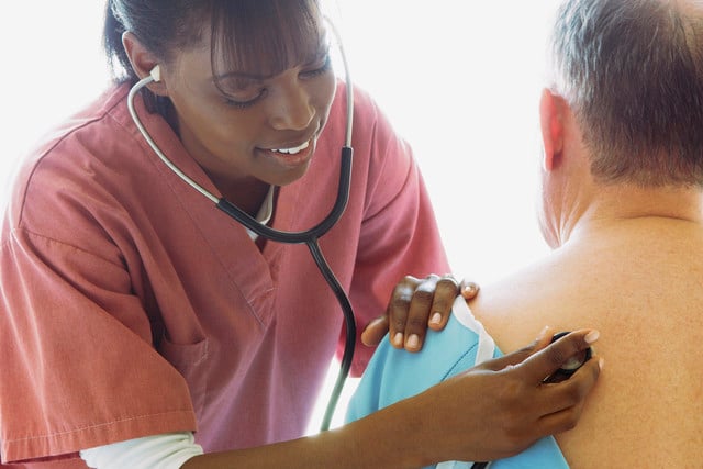 A healthcare professional in a pink uniform using a stethoscope on a patient’s back during a medical examination.