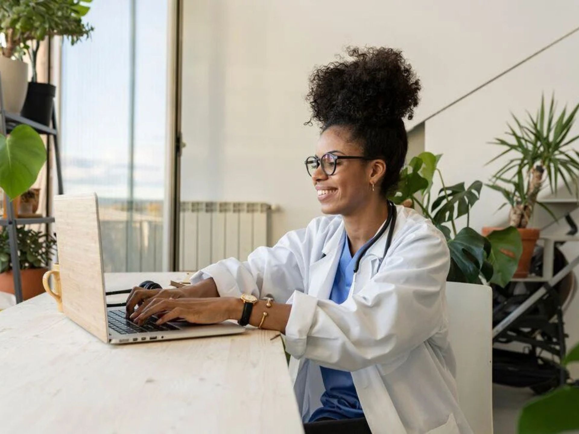 A medical professional in a white coat works on a laptop at a bright, plant-filled workspace, focused on their task.