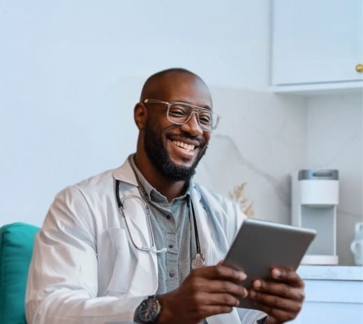 A smiling black man in a white coat is using a tablet, appearing engaged and professional in a clinical setting.