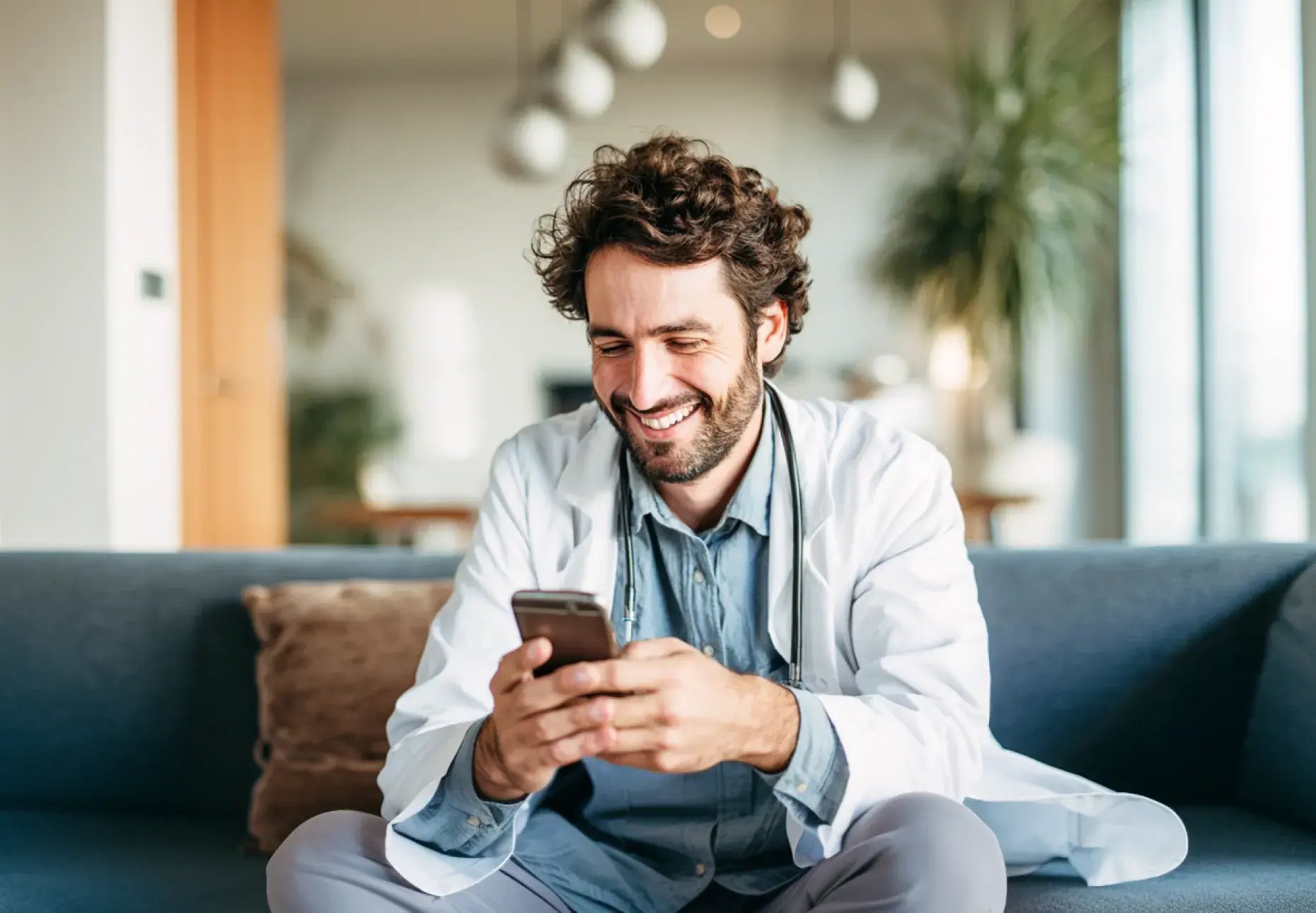 A male doctor in a white coat sits on a sofa, focused on his smartphone, with a stethoscope around his neck in a modern setting.
