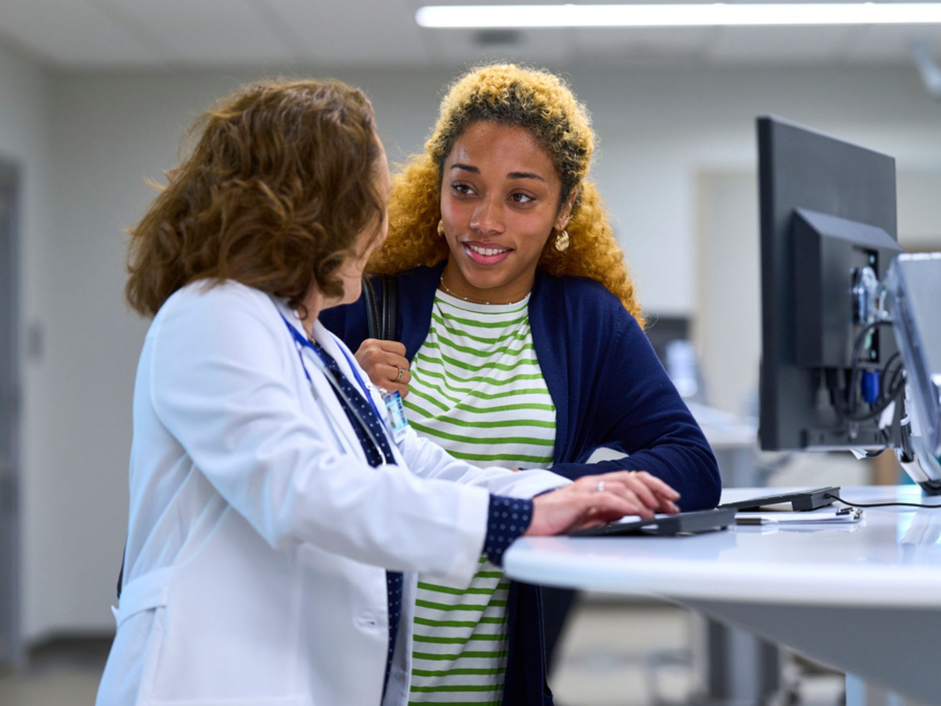 A healthcare professional in a lab coat discusses with a patient at a modern medical facility with computer equipment nearby.