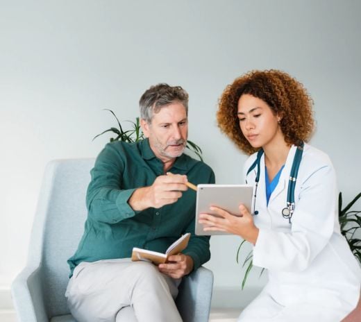 A doctor and patient sit on a couch, both focused on a tablet in their hands, discussing medical information.