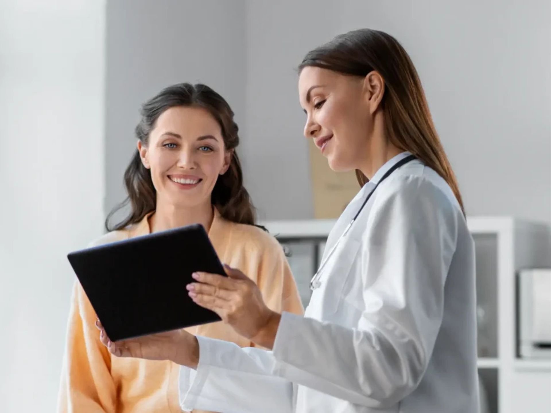 A doctor in a white coat discusses medical information with a patient, using a tablet in a bright, modern office setting.