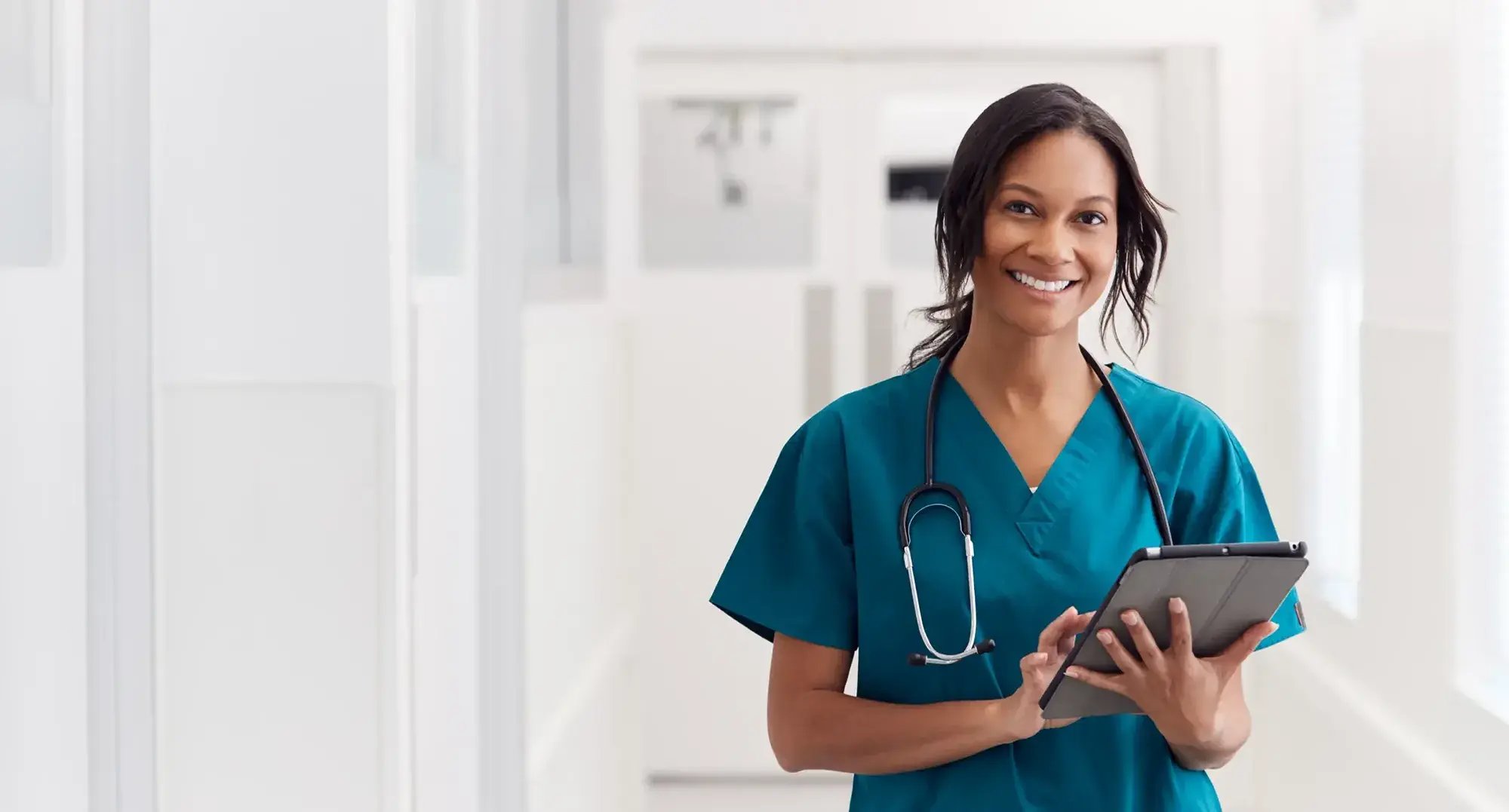 Portrait Of Smiling Female Doctor Wearing Scrubs In Hospital Corridor Holding Digital Tablet