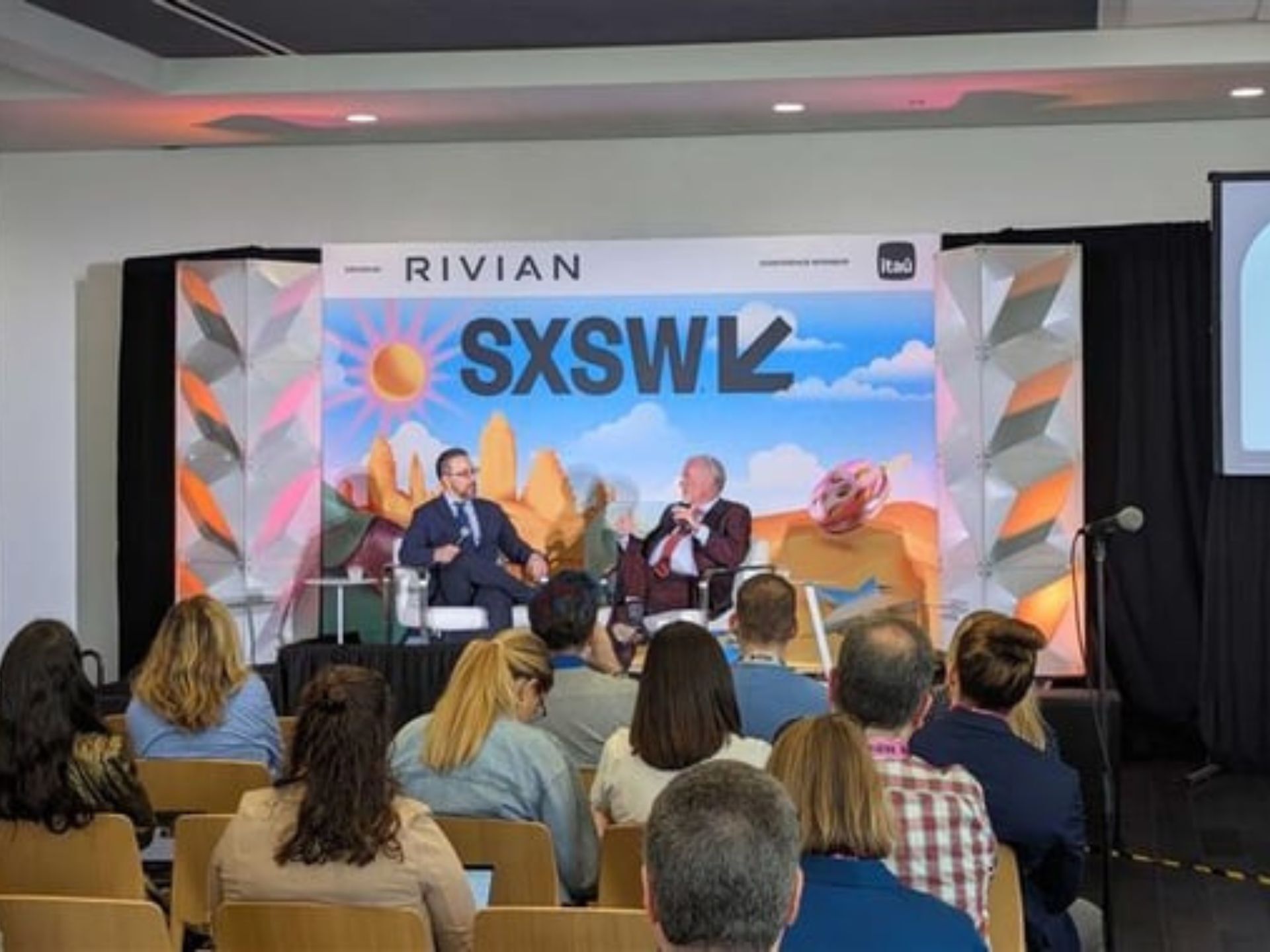 A conference panel discussion at SXSW, featuring two speakers on stage, with an audience engaged in the background and a colorful backdrop.