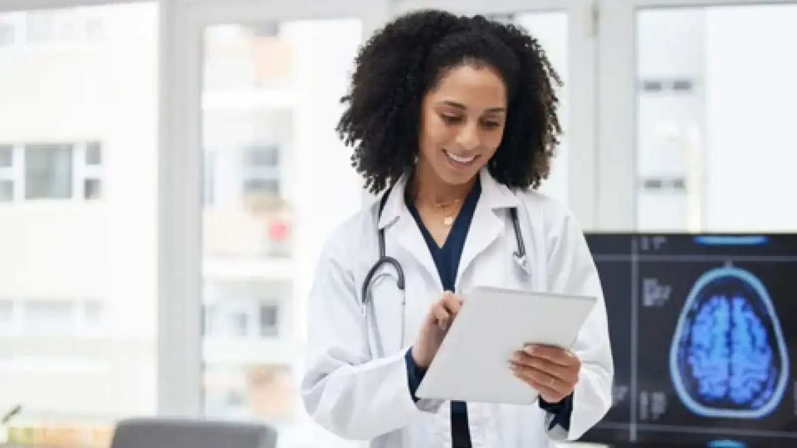 Woman doctor standing in front of a computer monitor reviewing notes on a clipboard.